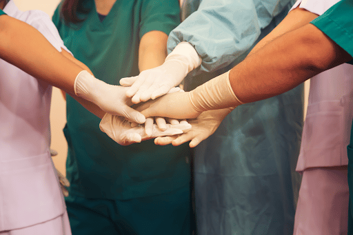 A group of healthcare professionals put their gloved hands together in a show of teamwork. Their faces can not be seen.