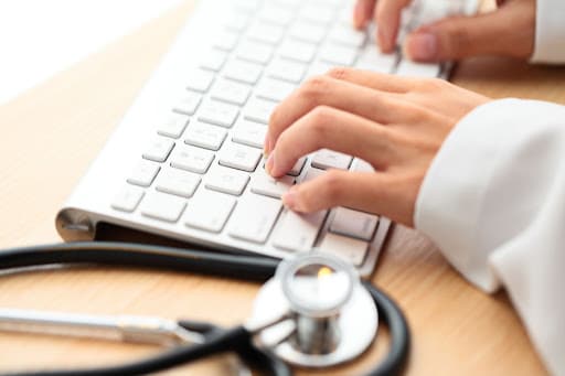 A woman types at a keyboard. She is a healthcare professional. Her face cannot be seen. The desk is wood.