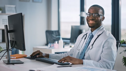 A healthcare professional sits at a desk. He is wearing glasses and smiling as he goes over his healthcare work. He has a blue tie.