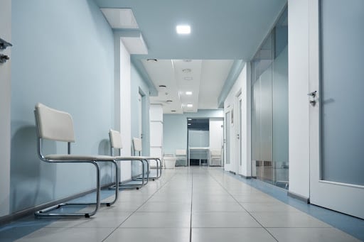 An empty hospital hallway is shown. The walls are baby blue. There are chairs sitting along the left hand wall. Doors are shown on the right.